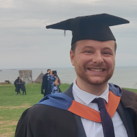 Photograph of a smiling man in graduation gown and mortarboard by the seaside.